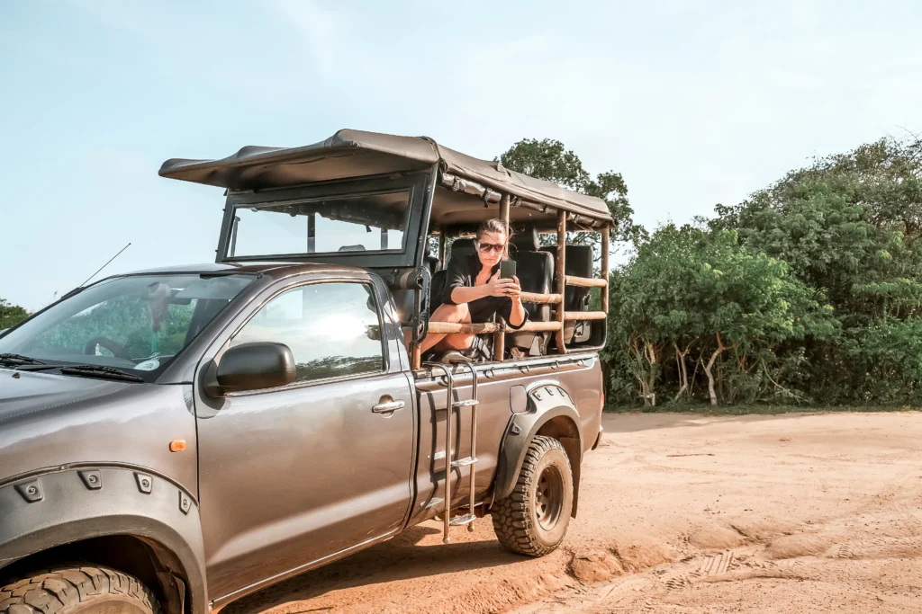 A woman is sitting in the back of a safari jeep, holding a phone, on a dusty path with green trees in the background. This image represents a wildlife safari experience offered by Ceylon Roots, a premier Tour Operator in Sri Lanka.