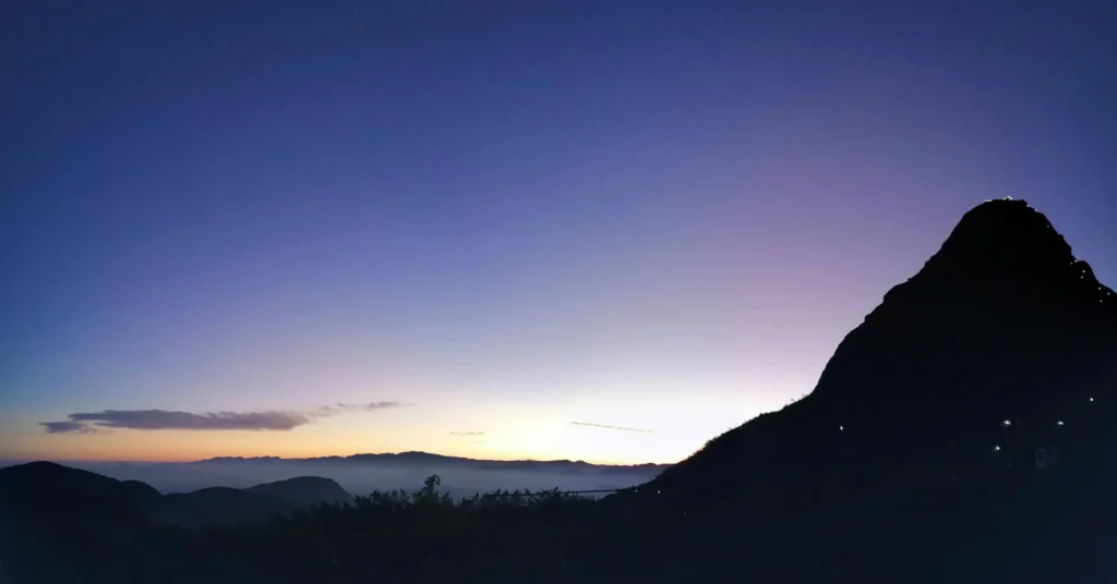 The silhouette of Adam's Peak (Sri Pada) against a colorful dawn sky, with a visible trail of lights from pilgrims ascending the mountain. This iconic pilgrimage and hiking experience is a popular offering from expert Tour Operators in Sri Lanka and a professional Tour Company in Sri Lanka.
