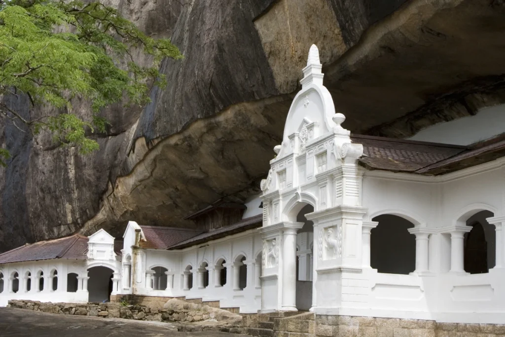 A white colonial-style building at the entrance of the Dambulla Cave Temple, built under a massive overhanging rock cliff. This UNESCO World Heritage site is a key cultural attraction and a highlight for any traveler on a tour with a leading Tour Company in Sri Lanka and professional Tour Operators in Sri Lanka.