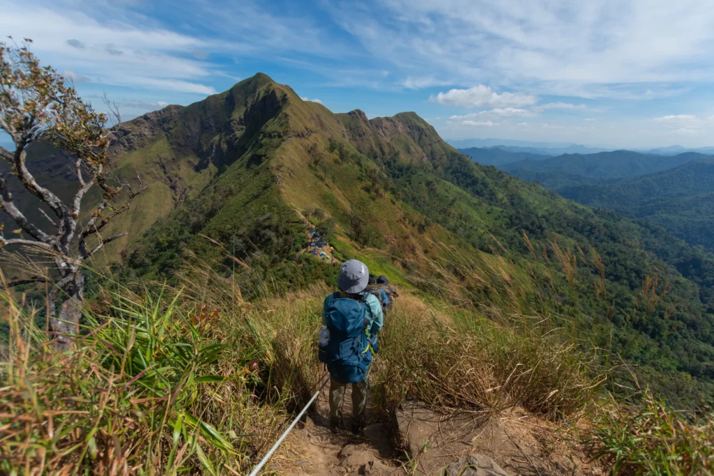 A woman with a backpack stands on a narrow mountain trail, looking out at a series of lush green peaks and valleys under a cloudy sky.