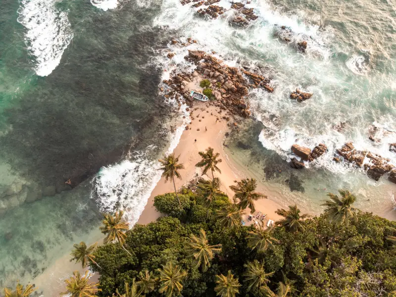 An aerial view captures a secluded tropical beach with rocky outcrops, crashing waves, and lush palm trees, showcasing the stunning coastal scenery often featured in tours offered by top-rated Tour Operators in Sri Lanka and a reliable Tour Company in Sri Lanka.