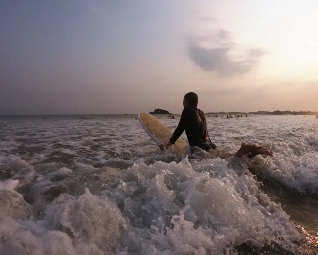 A surfer with a board wades into the waves at sunset, with other surfers visible in the distance. This is a popular activity offered by expert Tour Operators in Sri Lanka for those seeking beach and water sports experiences with a professional Tour Company in Sri Lanka.