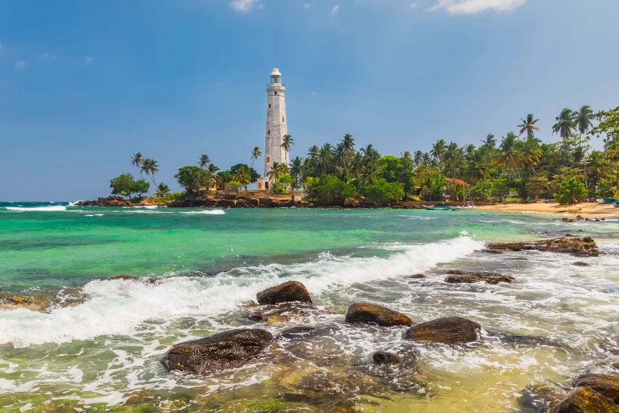 A white lighthouse stands on a rocky outcrop surrounded by palm trees, with waves crashing on a sandy beach. This scenic coastal landmark is a popular attraction on tours organized by a top Tour Company in Sri Lanka and expert Tour Operators in Sri Lanka.